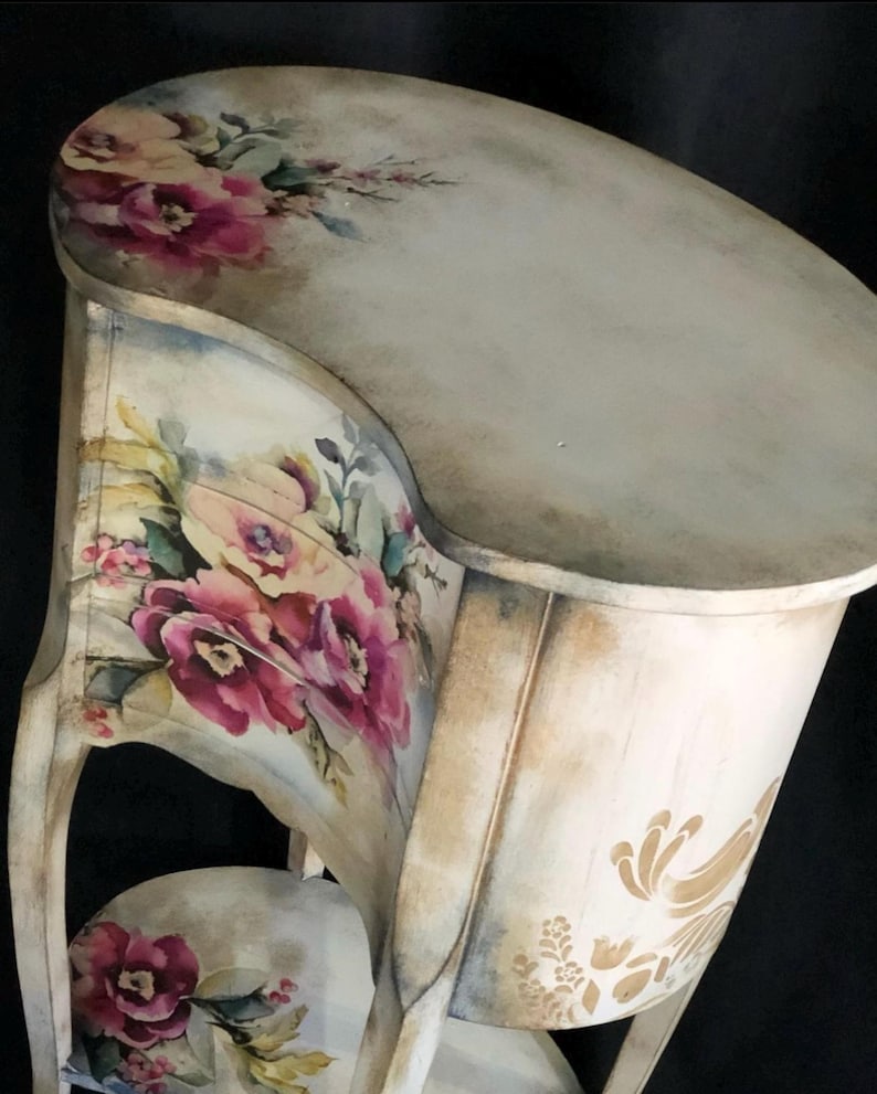 May include: A white painted wooden side table with a curved top and a floral design. The table has a lower shelf and a floral design on the front. The table is distressed and has a vintage look.