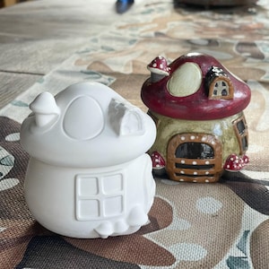 Peut inclure: Deux maisons en forme de champignon en céramique, l'une peinte en rouge, marron et vert avec des pois blancs, l'autre en blanc avec une fenêtre. Les deux maisons ont un toit en forme de champignon.
