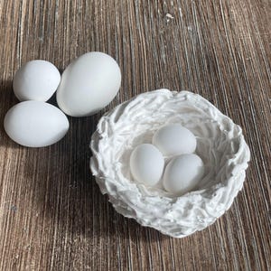 Op de afbeelding: Een wit keramisch vogelnest met drie witte eieren erin. Het nest ligt op een houten oppervlak.