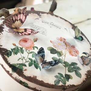 May include: A round, white box with a distressed brown edge. The box is decorated with a floral design featuring pink roses and green leaves. There are several butterflies on the box, including a pink and white butterfly, a blue and black butterfly, and a yellow and brown butterfly. The text "Rose Garden" is printed on the box.
