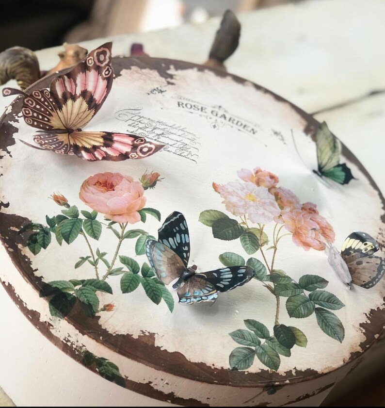 May include: A round, white box with a distressed brown edge. The box is decorated with a floral design featuring pink roses and green leaves. There are several butterflies on the box, including a pink and white butterfly, a blue and black butterfly, and a yellow and brown butterfly. The text "Rose Garden" is printed on the box.