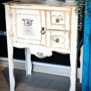 Peut inclure: Une table d'appoint blanche vieillie avec deux tiroirs et une porte de placard. La porte du placard est orn&eacute;e d'un motif floral d&eacute;coratif et du texte "Hello Strangers" imprim&eacute; dessus. Les tiroirs sont munis de poign&eacute;es m&eacute;talliques d&eacute;coratives.