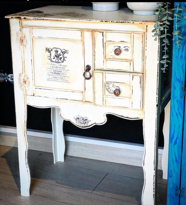 Peut inclure: Une table d'appoint blanche vieillie avec deux tiroirs et une porte de placard. La porte du placard est orn&eacute;e d'un motif floral d&eacute;coratif et du texte "Hello Strangers" imprim&eacute; dessus. Les tiroirs sont munis de poign&eacute;es m&eacute;talliques d&eacute;coratives.