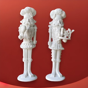 May include: Two white ceramic nutcracker figurines. One holds a ball and the other holds a tray with a snowflake design.