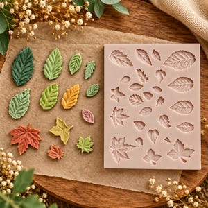 May include: A rectangular silicone mold featuring various leaf-shaped cavities, accompanied by an assortment of small, colorful, molded leaf shapes in shades of green, yellow, orange, and pink, arranged on a piece of brown parchment paper.