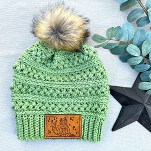 May include: Green crocheted beanie with a brown leather patch featuring a snail and mushrooms. The beanie has a fluffy brown pom pom on top.