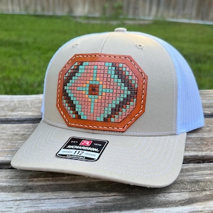 May include: A beige and white trucker hat with a brown leather patch featuring a geometric pattern in shades of pink, turquoise, and brown. The patch has a stitched border and the hat has an adjustable strap with the text "Richardson 112 Trucker Adjustable" printed on it.