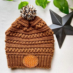 May include: A brown crocheted beanie with a brown pom pom on top and a brown leather waffle patch on the front.