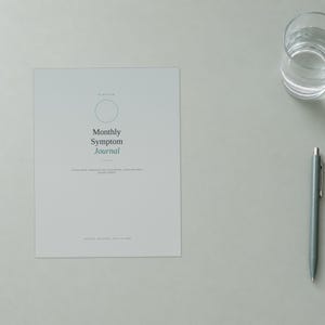 May include: A light gray Monthly Symptom Journal with a thin circular outline, dark gray text, and a green accent word. The journal cover features the title Monthly Symptom Journal and smaller descriptive text. A gray pen sits to the right.