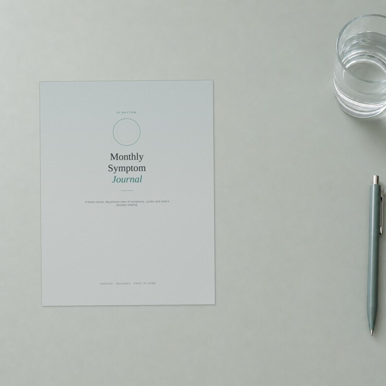 May include: A light gray Monthly Symptom Journal with a thin circular outline, dark gray text, and a green accent word. The journal cover features the title Monthly Symptom Journal and smaller descriptive text. A gray pen sits to the right.