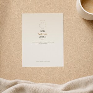 May include: A rectangular paper journal cover in a light cream colour featuring a thin circular outline, the text RSD Reflection Journal, and smaller printed text below. The design is centred on the page with a clean, minimalist layout.