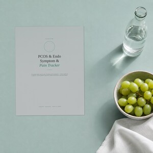 May include: A light grey rectangular symptom tracker document with the text PCOS & Endo Symptom & Pain Tracker printed in black and green. The document is positioned next to a clear glass water bottle and a small bowl filled with green grapes.