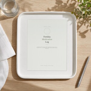 Op de afbeelding: Een wit rechthoekig dienblad met een geprint document getiteld Fertility Medication Log. Het papier bevat een cirkelvormig icoon, beschrijvende tekst en de voettekst Hopeful Steps, Reusable, Print at Home op een lichte achtergrond.