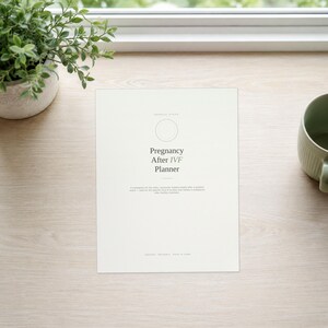 May include: A white planner cover with the text Pregnancy After IVF Planner, a circular outline, and smaller text below. The cover features a clean, minimalist design with dark grey lettering on a light background, centred on a wooden surface.