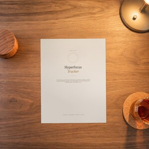 May include: A white rectangular paper sheet titled Hyperfocus Tracker with a circular outline at the top. The text includes a description of focus and the words Created, Fillable, Print at Home. The paper is centred on a dark wood surface.