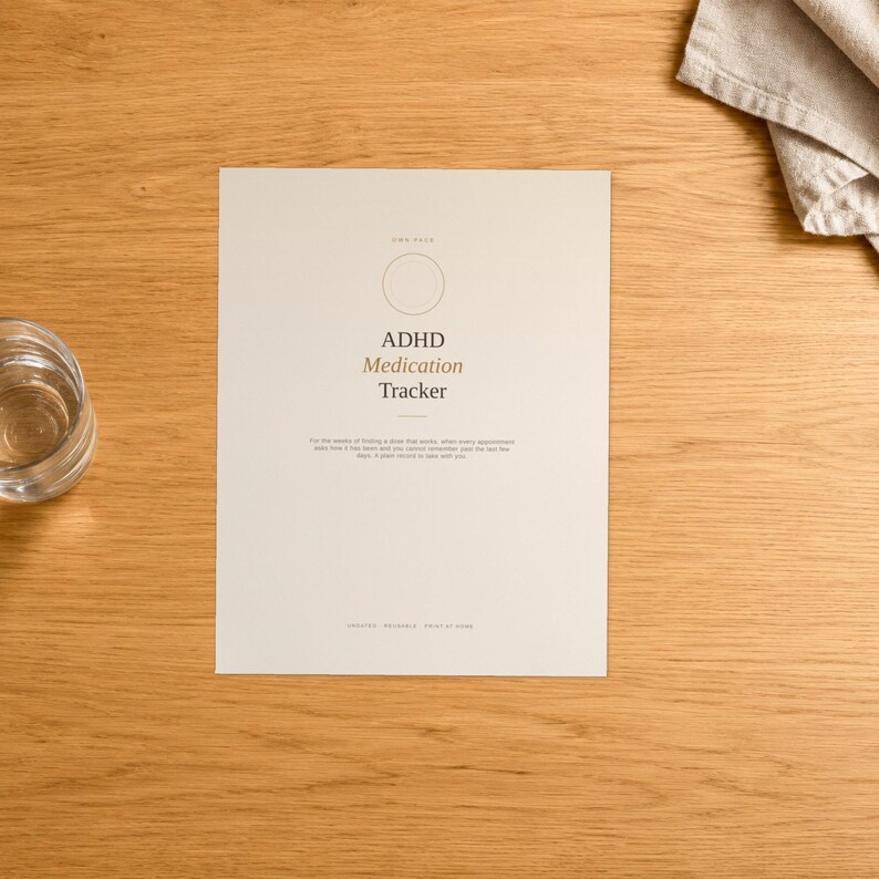 K&ouml;nnte beinhalten: Ein cremefarbenes ADHD Medication Tracker Papierblatt mit einem kreisf&ouml;rmigen Umriss oben, zentriertem schwarzem und braunem Text sowie einem kurzen Anweisungsabsatz unter dem Titel. Das Dokument liegt auf einer hellen Holzoberfl&auml;che.