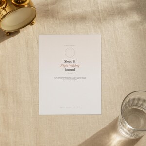 May include: A rectangular white journal cover with the text Sleep & Night Waking Journal printed in black and red. The cover features a thin circular outline above the title and smaller text below, positioned near a gold alarm clock and a glass.