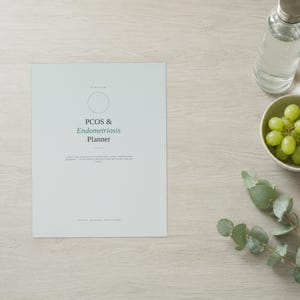 May include: A light grey planner with a circular outline and the text PCOS & Endometriosis Planner printed in dark grey and green. The cover includes a subtitle and small text at the bottom, presented in a clean, minimalist layout on a flat surface.