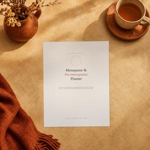 May include: A white planner cover with the text Menopause & Perimenopause Planner in black and pink font. The cover features a thin circle outline above the title and smaller text below. It is positioned near a brown ceramic vase and a tea cup.