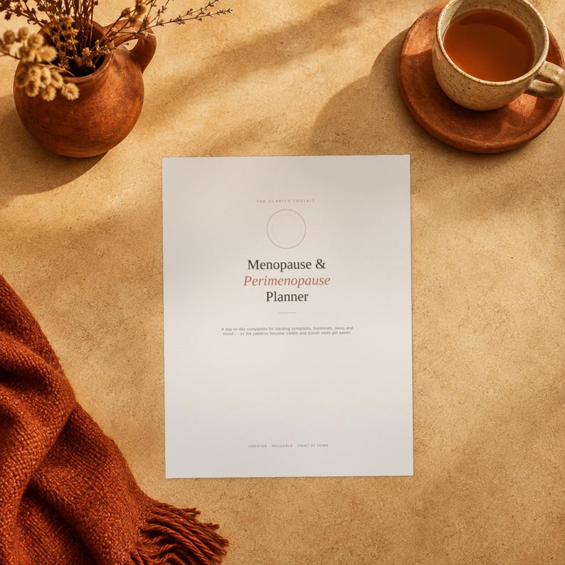 May include: A white planner cover with the text Menopause & Perimenopause Planner in black and pink font. The cover features a thin circle outline above the title and smaller text below. It is positioned near a brown ceramic vase and a tea cup.