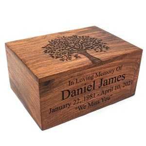 Puede incluir: Una urna de cremación de madera con un diseño de árbol grabado en la tapa. La urna está personalizada con el texto "In Loving Memory Of Daniel James January 22, 1981 - April 10, 2021 "We Miss You""