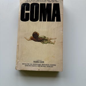 Può includere: Una copertina di un libro tascabile con il titolo "COMA" in grassetto in lettere nere. La copertina presenta un dipinto di una persona che galleggia nell'aria, circondata da corde. Il libro è di Robin Cook ed è un thriller medico.