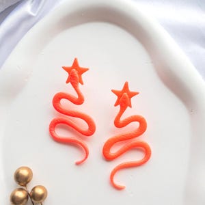 Könnte beinhalten: Ein Paar leuchtend orangefarbene Ohrringe. Jeder Ohrring hat einen Stern an der Spitze und ein schlangenartiges Design. Die Ohrringe werden auf einem weißen, oval geformten Teller präsentiert. Zusätzliche goldfarbene Zierpins sind sichtbar.