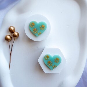 May include: Two white, geometric-shaped objects with teal heart-shaped designs. Each heart has gold paw prints. One object is round, the other is hexagonal. Gold decorative spheres on stems are to the left.