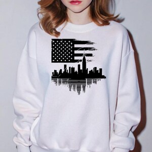May include: White sweatshirt with a black graphic print of the New York City skyline with the American flag in the background.