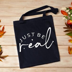 May include: A black canvas tote bag with white text that reads "Just Be Real" with a small heart and stars around the text.