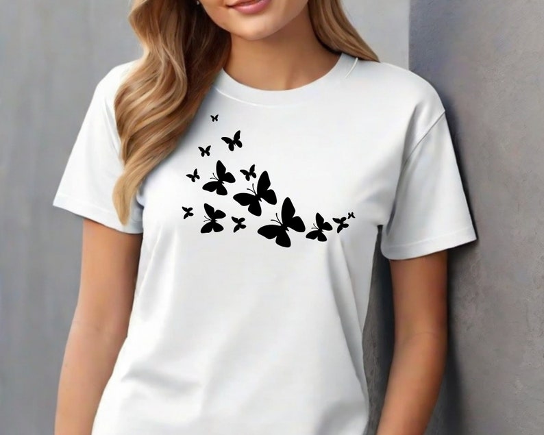 Puede incluir: Camiseta blanca con una silueta negra de una bandada de mariposas volando en diagonal.