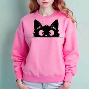 K&ouml;nnte beinhalten: Ein rosa Sweatshirt mit einer schwarzen Katze, die &uuml;ber den oberen Rand schaut. Die Katze hat gro&szlig;e Augen und einen verspielten Ausdruck.