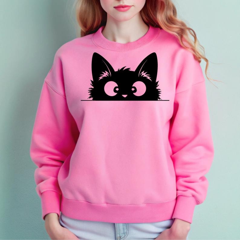 K&ouml;nnte beinhalten: Ein rosa Sweatshirt mit einer schwarzen Katze, die &uuml;ber den oberen Rand schaut. Die Katze hat gro&szlig;e Augen und einen verspielten Ausdruck.