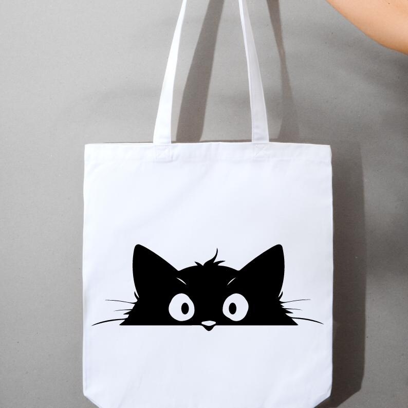 K&ouml;nnte beinhalten: Wei&szlig;e Canvas-Tragetasche mit einer schwarzen Silhouette einer Katze, die &uuml;ber den Rand schaut. Die Katze hat gro&szlig;e Augen und eine kleine Nase.