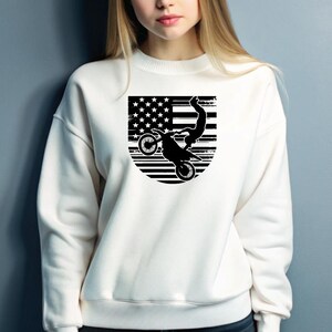 May include: White sweatshirt with a black graphic of a person performing a stunt on a dirt bike in front of an American flag.