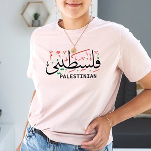 May include: A pink t-shirt with the Arabic word for "Palestinian" in red, green, and black, along with the word "Palestinian" in English below it.