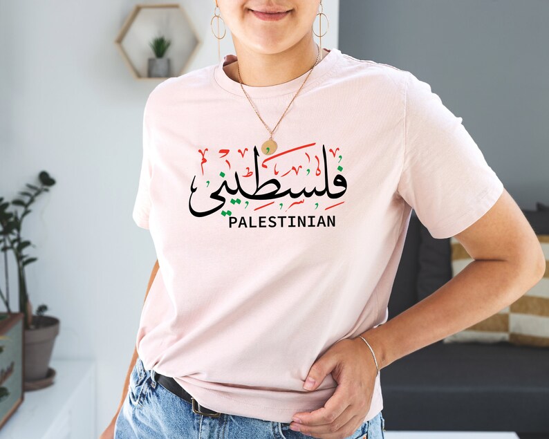 May include: A pink t-shirt with the Arabic word for "Palestinian" in red, green, and black, along with the word "Palestinian" in English below it.