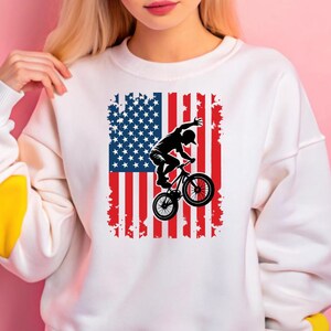May include: White sweatshirt with a graphic of a person riding a BMX bike in front of an American flag.