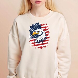 May include: A white sweatshirt with a distressed American flag design featuring a bald eagle. The eagle's head is facing the left side of the image. The flag is red, white, and blue.