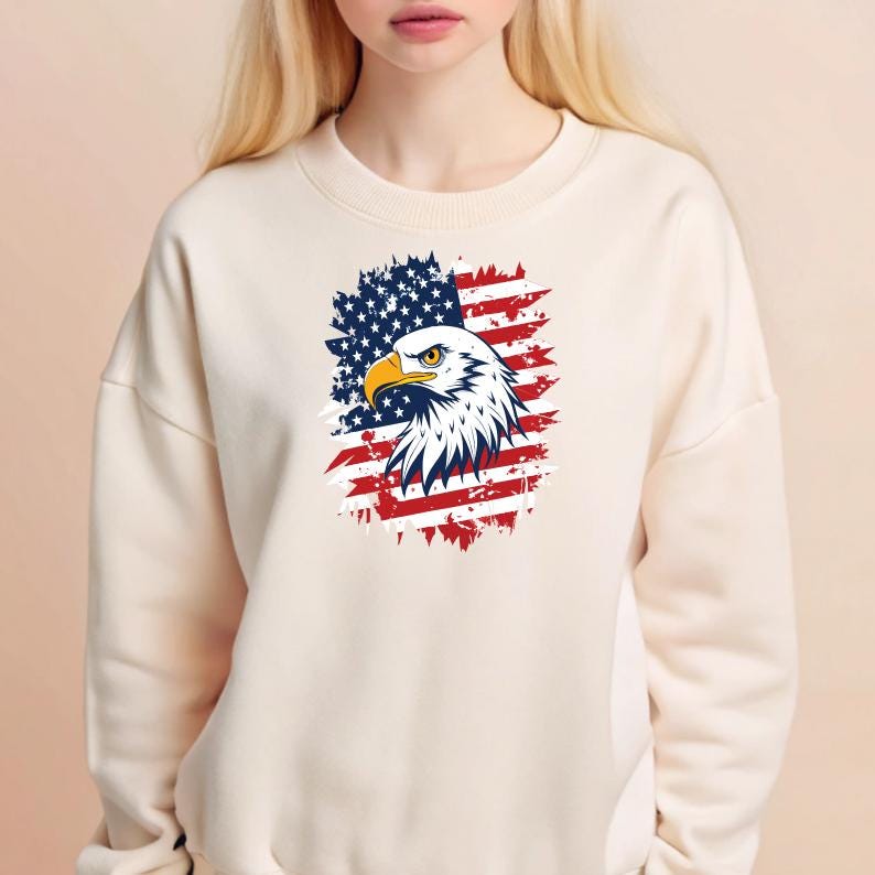 May include: A white sweatshirt with a distressed American flag design featuring a bald eagle. The eagle's head is facing the left side of the image. The flag is red, white, and blue.