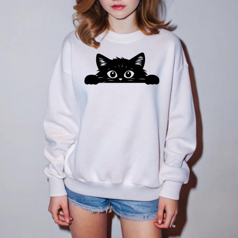 K&ouml;nnte beinhalten: Wei&szlig;er Pullover mit einer schwarzen Katze, die &uuml;ber den Rand schaut. Die Katze hat gro&szlig;e Augen und ein schlichtes Design.