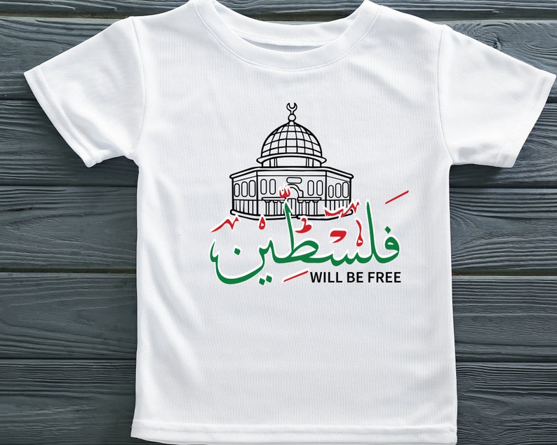 May include: White t-shirt with a black line drawing of the Dome of the Rock, a mosque in Jerusalem. The Arabic text "فلسطين" (Palestine) is written in green and red, and the English text "WILL BE FREE" is written in black below.