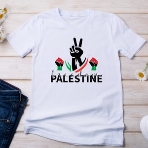 May include: White t-shirt with a black peace sign and the words "freedom PALESTINE" in black and green. There are two red fists on either side of the peace sign.