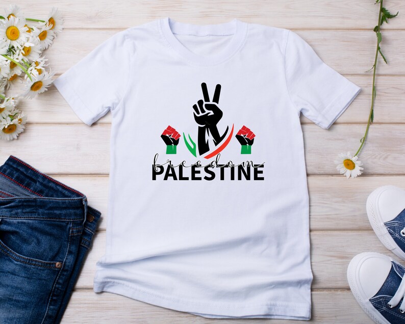 May include: White t-shirt with a black peace sign and the words "freedom PALESTINE" in black and green. There are two red fists on either side of the peace sign.