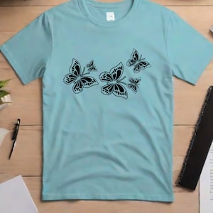Puede incluir: Una camiseta azul claro con un dise&ntilde;o de mariposa negra. Las mariposas son de diferentes tama&ntilde;os y est&aacute;n agrupadas en la parte delantera de la camiseta.