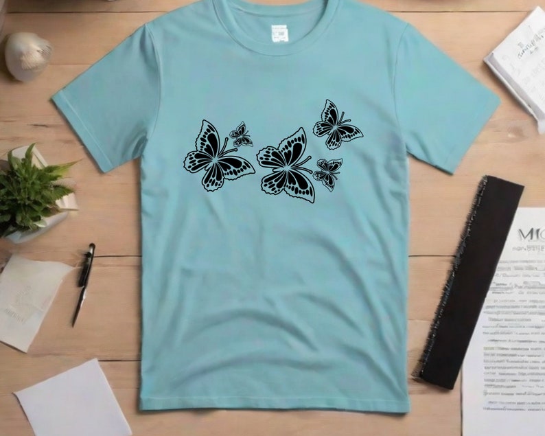 Puede incluir: Una camiseta azul claro con un dise&ntilde;o de mariposa negra. Las mariposas son de diferentes tama&ntilde;os y est&aacute;n agrupadas en la parte delantera de la camiseta.