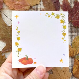 Op de afbeelding: Een vierkant wit papieren briefje met een decoratieve rand met geelgroene rankillustraties langs de linker- en bovenrand. Een kleine oranje pompoen staat afgebeeld in de linkerbenedenhoek, geaccentueerd door kleine gele en roze sterretjes.