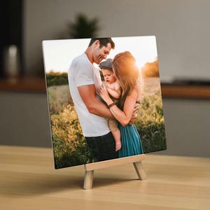 May include: A square photo print on a small wooden easel. The photo shows a family of three, a man, a woman, and a baby, in a field of tall grass. The family is embracing. The photo is displayed on a wooden easel.