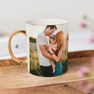 May include: White ceramic mug with a gold rim and handle. The mug features a photo of a family of three in a field. The family is made up of a man, a woman, and a young child.