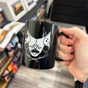 Könnte beinhalten: Schwarze Keramiktasse mit einem weißen Motiv einer Fledermaus mit Totenkopf und Knochen.